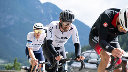 Die drei Rennradfahrer der Spitzengruppe fahren konzentriert. Es sind nur die Oberkörper und Köpfe zu sehen. Im Hintegrund ist ein Berg zu erkennen.