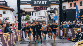Athleten starten den Hindernislauf durch einen hölzernen Zielbogen. Sie tragen spotliche Sachen. Der Himmel ist bewölkt.