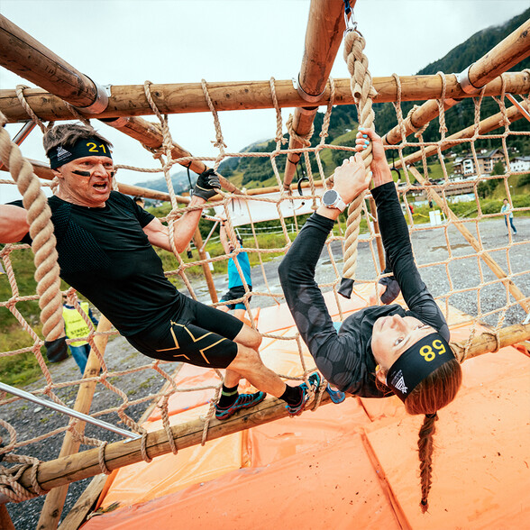 Teilnehmerin hängt kopfüber an einem Hindernis und hangelt sich entlang der Taue entlang. Ein Mann daneben blickt begeistert und erstaunt drauf.
