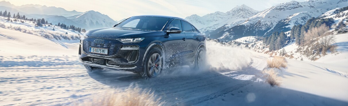Audi electric car races through a snowy, alpine country road on a beautiful, slightly cloudy winter day.