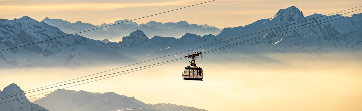 Die Vallugabahn 1 fährt bergauf vor einem winterlich Alpenpanorama im Nebelmeer.