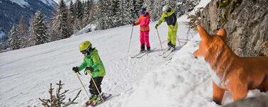 Kind und Eltern fahren auf einer flachen Skipiste an einer Fuchs-Figur vorbei.