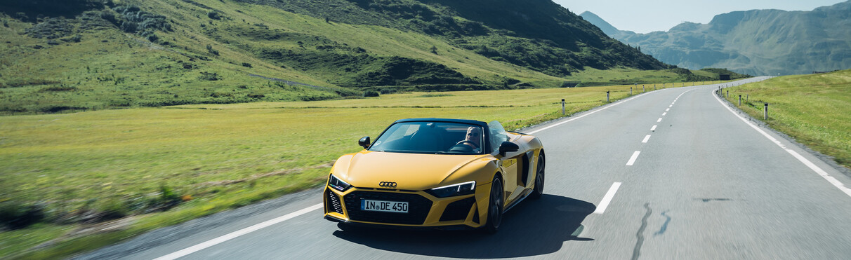 Gelber Audi R8 fährt eine hochalpine Freilandstraße entlang. Im Sommer bei schönem Wetter.