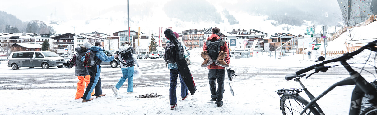 Zwei Wintersportler von hinten abgelichtet, wie sie den Bahnhof St. Anton am Arlberg verlassen, im Hintergrund sieht man das verschneite Dorf.