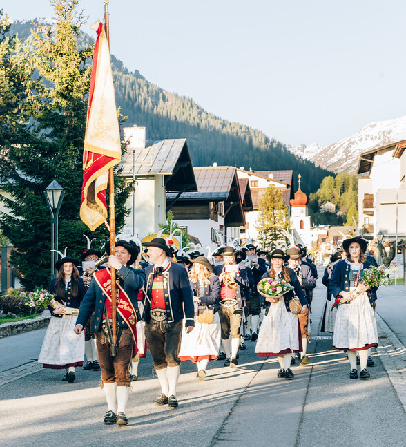 Die Musikkapelle St. Anton wie sie durch die Gassen des Dorfes marschieren in blauer traditioneller Trachten-Kleidung.