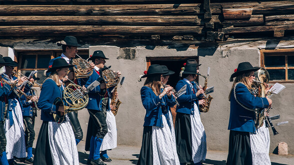 Musikkapelle Pettneu marschiert an einem holzernen Stadl vorbei in Tracht. Von der Seite fotografiert.