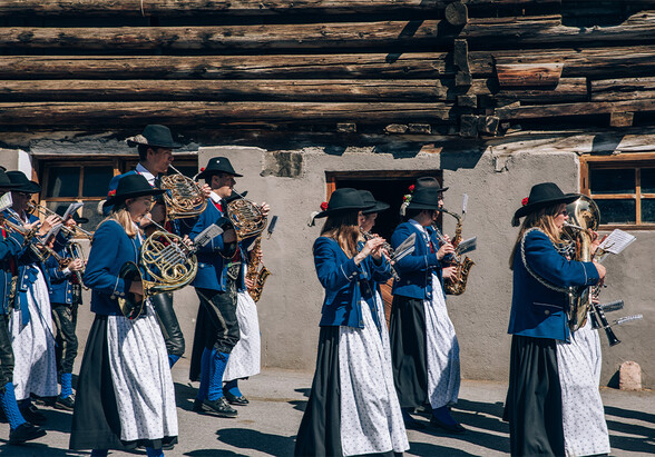 The Pettneu marching band marches past a wooden barn in traditional costume. Photographed from the side.