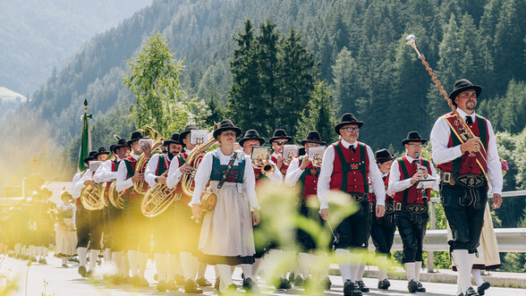 Musikkapelle Strengen marschiert über ein Freilandstraße, im hintegrund sieht man die waldigen Berge des Stanzertals.