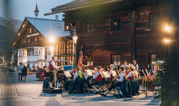 Musikkapelle St. Jakob spielt ein Konzert im Freien vor einem Holzhaus.
