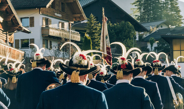 Eine Musikkapelle zieht in traditioneller Tracht durch ein Dorf, gesehen von hinten, begleitet von Fahnen und Wohnhäusern.