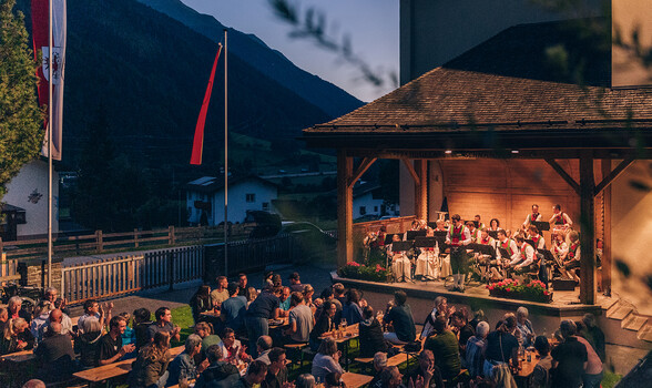 Eine Musikkapelle bewegt sich musizierend durch ein alpines Dorf mit Fahnen, Häusern und Bergkulisse.