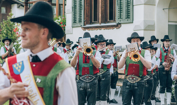 Eine Musikkapelle zieht durch eine enge Gasse, angeführt von einer Fahne, umgeben von Häusern und Kirche.