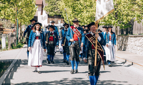 Eine Musikkapelle marschiert in geordneter Formation durch eine schmale Dorfstraße und spielt Blasinstrumente.