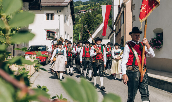 Eine Musikkapelle bewegt sich musizierend durch ein alpines Dorf mit Fahnen, Häusern und Bergkulisse.