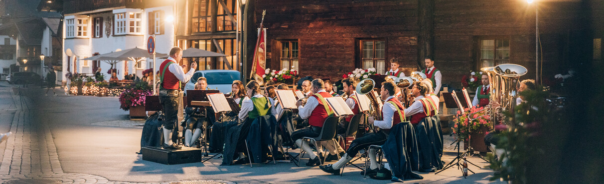 Eine Musikkapelle spielt abends auf einem Dorfplatz vor historischen Holzhäusern, geleitet von einem Dirigenten.