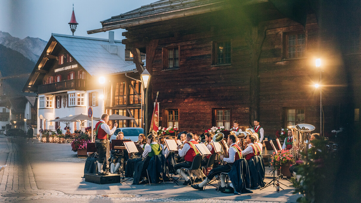 A band plays in the evening on a village square in front of historic wooden houses, led by a conductor.