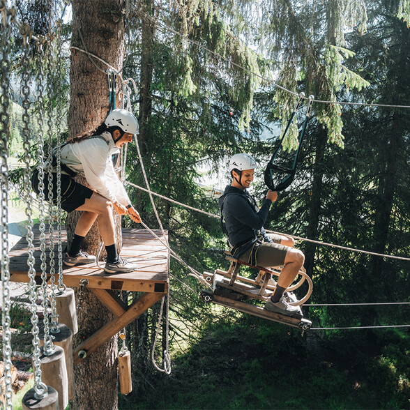 Persona assicurata che percorre una teleferica nel percorso avventura tra gli alberi, mentre un'altra persona aspetta su una piattaforma.