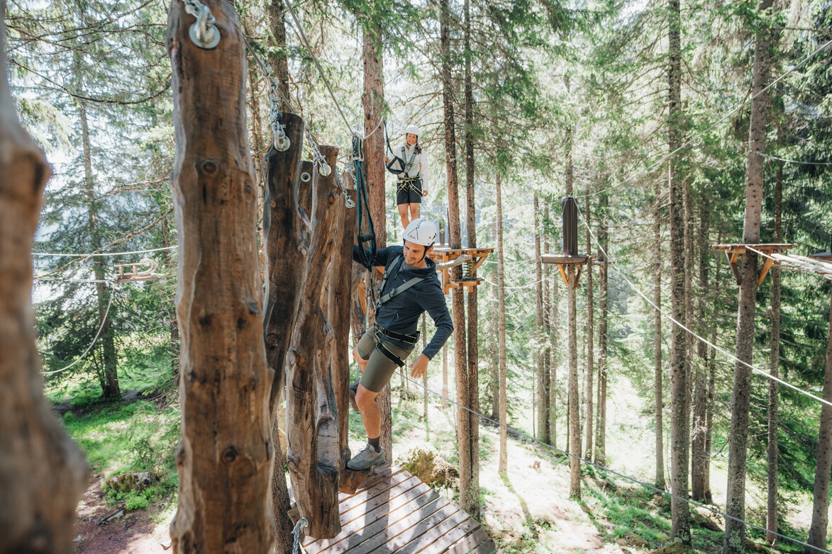 Holzhindernis im Waldseilgarten Gesicherte Person balanciert über ein Holzhindernis im Waldseilgarten, während eine weitere Person im Hintergrund wartet.