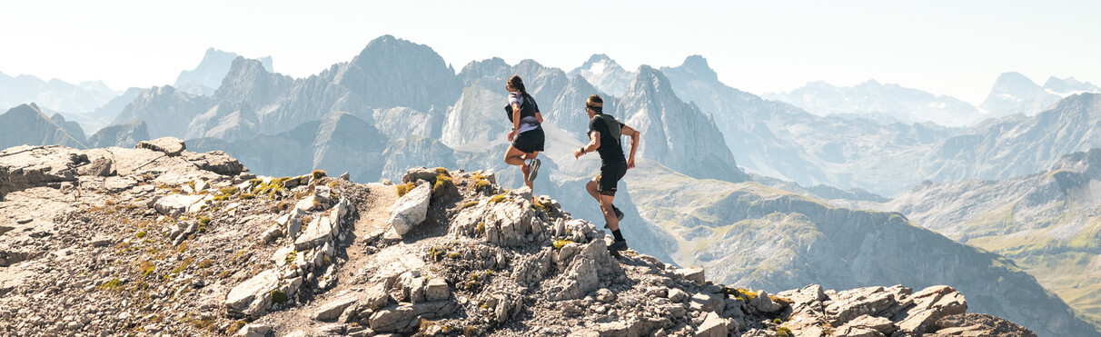 Two trail runners run along a rocky mountain ridge with an impressive Alpine panorama in the background.