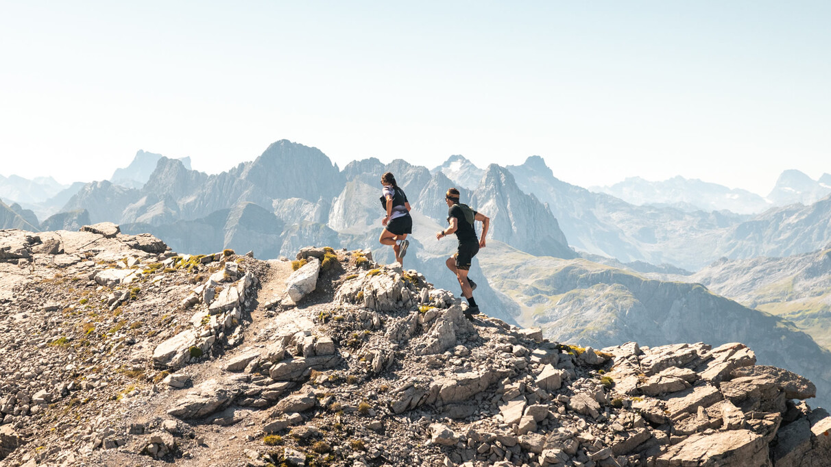 Two trail runners run along a rocky mountain ridge with an impressive Alpine panorama in the background.