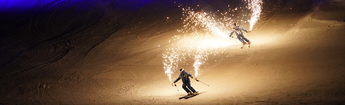 Zwei Skifahrer mit Funken-Fackeln gleiten bei Nacht über eine Piste.
