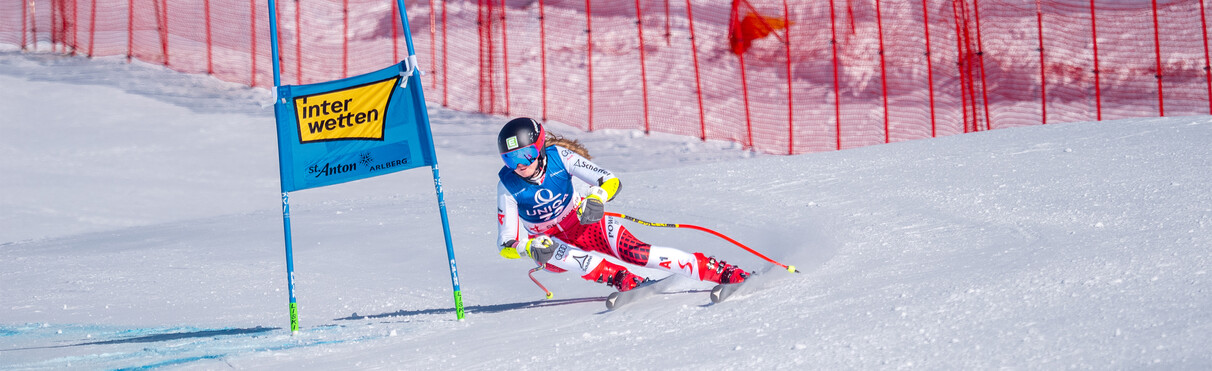 Skirennläuferin in rot-weißer Ausrüstung fährt an einem blauen Tor auf einer schneebedeckten Piste vorbei