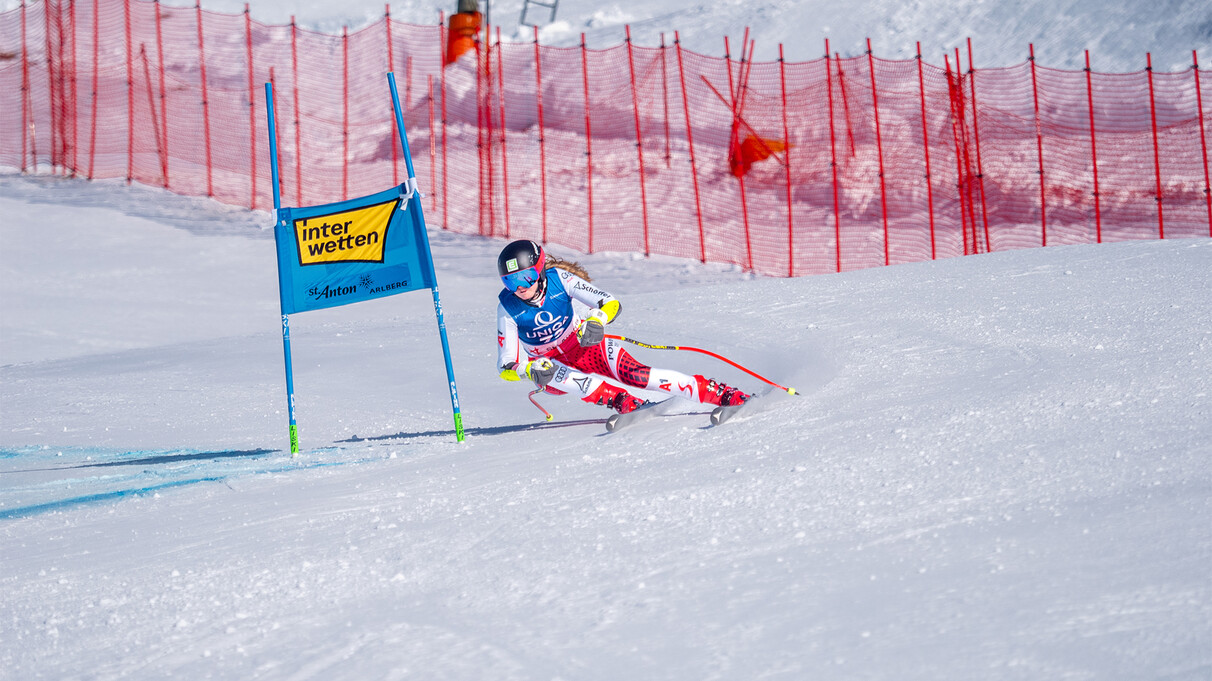 Skirennläuferin in rot-weißer Ausrüstung fährt an einem blauen Tor auf einer schneebedeckten Piste vorbei