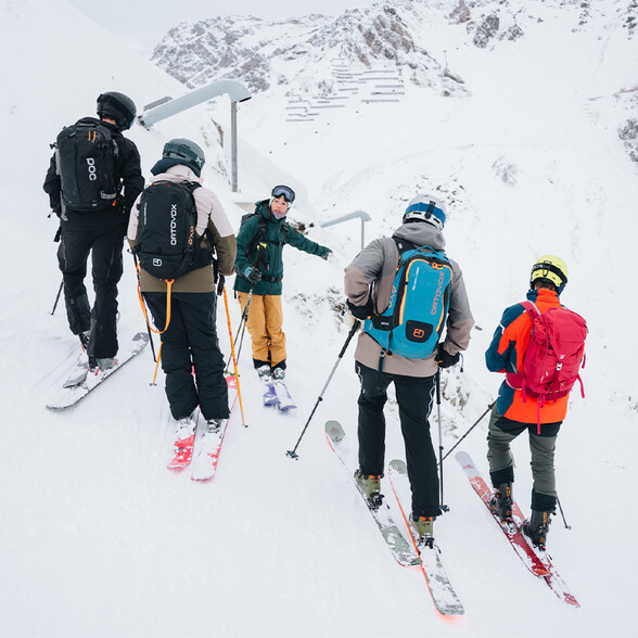 Group of skiers standing on snow facing a mountain with one person pointing ahead