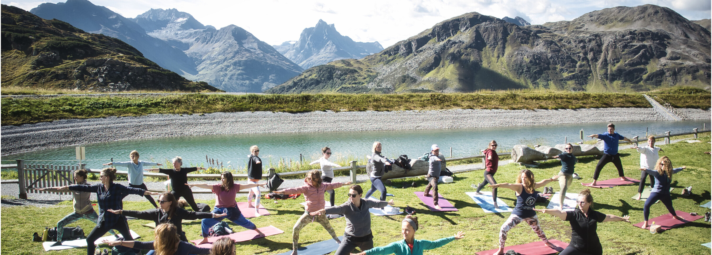 Yoga in St. Anton am Arlberg Gruppe von Menschen praktiziert Yoga auf bunten Matten auf einer Wiese vor einem Bergsee mit dem Patteriol im Hintergrund