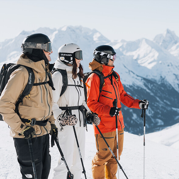 Drei Skifahrer mit Helmen, Skibrillen und Skistöcken stehen nebeneinander vor verschneiten Bergen