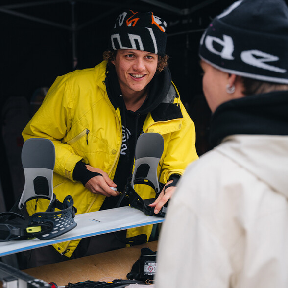 Junger Mann in gelber Jacke und schwarzer Mütze montiert Snowboardbindungen an einem Snowboard und spricht mit einer Person in weißer Jacke und schwarzer Mütze.