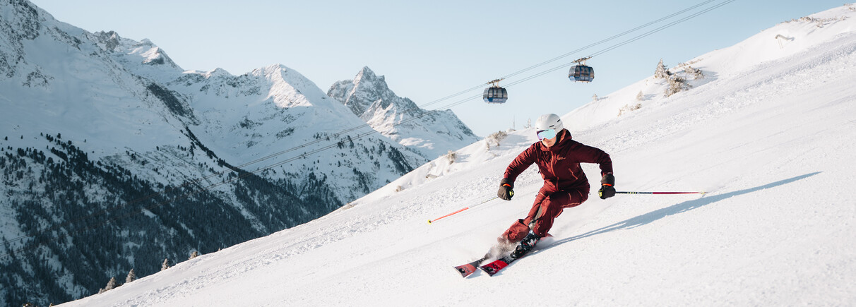 Skifahrer in roter Kleidung fährt auf schneebedeckter Piste mit Bergpanorama und Gondelbahnen im Hintergrund
