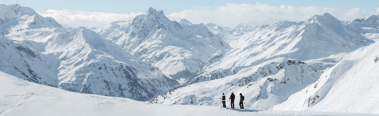 Three people with ski equipment stand on a snow-covered mountain peak with a range of snow-covered mountains in the background