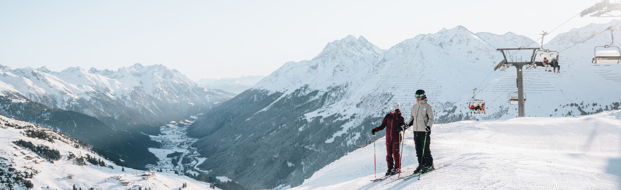 Two skiers standing on a snowy mountain slope overlooking a valley with snow-covered peaks and a ski lift with chairs