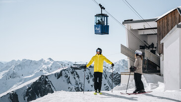 Prices and News Two skiers in helmets and ski gear stand near a cable car station with a blue cable car suspended above snowy mountain peaks