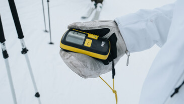 Safety Hand in a white winter jacket and glove holding a yellow and black handheld device with a screen and buttons in a snowy environment