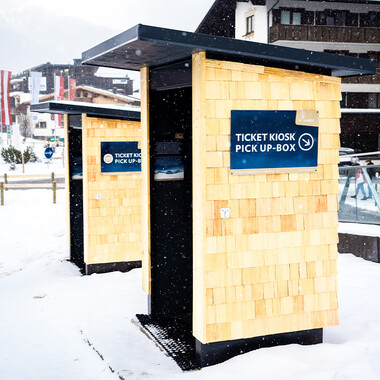 Two small wooden kiosks with shingles and blue signs reading 'TICKET KIOSK PICK UP-BOX' in the snow in front of buildings.