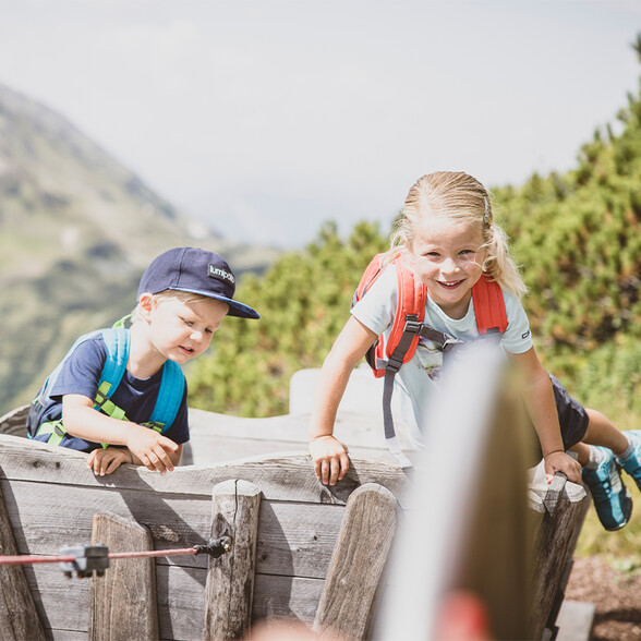 Zwei Kinder mit Rucksäcken klettern auf einem Holzwagen in einer Berglandschaft