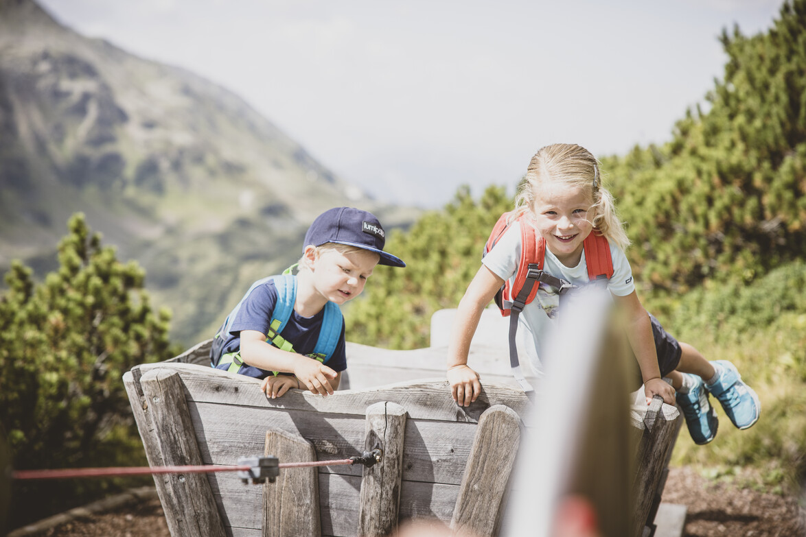Mutspuren Rundwanderweg am Galzig Zwei Kinder mit Rucksäcken klettern auf einem hölzernen Spielgerät in einer Berglandschaft