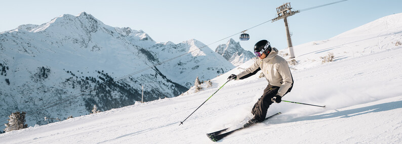 St. Anton am Arlberg – Winter Skier in beige jas en zwarte helm die een bocht maakt op een besneeuwde helling met bergtoppen op de achtergrond
