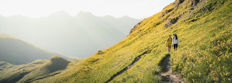 St. Anton am Arlberg – Zomer Twee wandelaars op een smal bergpad omgeven door groene hellingen met zonlicht en bergen op de achtergrond.