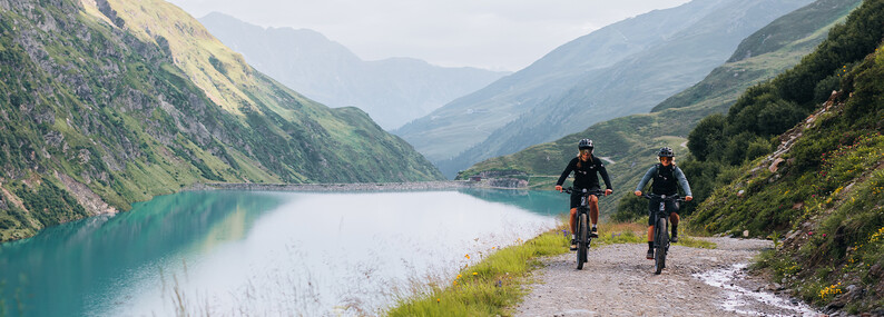 E-bike en mountainbike Twee mountainbikers rijden op een gravelpad langs een bergmeer met bergen op de achtergrond