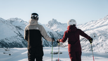 Ski Arlberg Twee skiërs met helmen en skistokken kijken uit over besneeuwde bergen in het skigebied Arlberg.