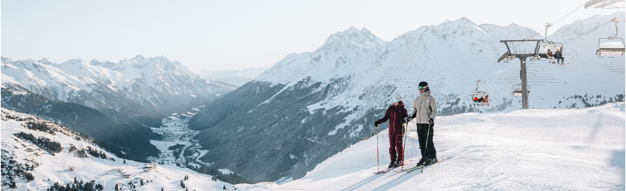 Ski Arlberg Twee skiërs staan op een besneeuwde bergtop met uitzicht op een dal en een stoeltjeslift rechtsboven.