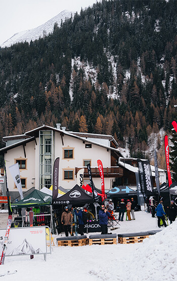 Several tents with brand logos and people in front of a multi-story building in a snowy mountain area with forested slopes in the background.