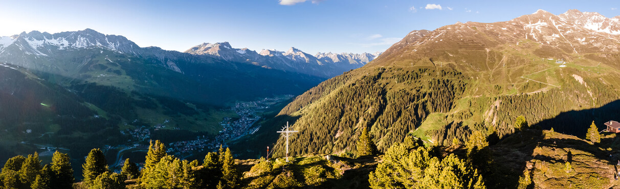 Panoramaaufnahme vom Wirt Gipfelkreuz mit Blick auf St. Anton am Arlberg, das sich sich zwischen den Bergketten zurecht findet. Diffuses Abendlicht.