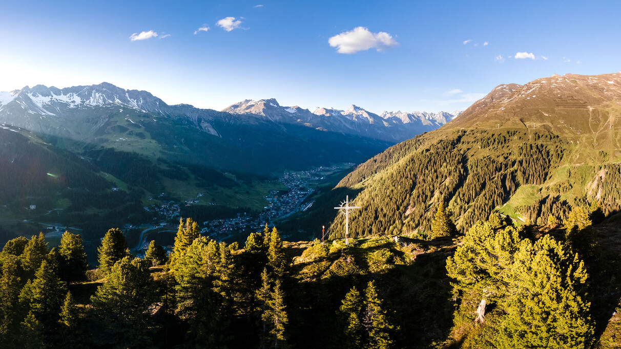 Panoramaaufnahme vom Wirt Gipfelkreuz mit Blick auf St. Anton am Arlberg, das sich sich zwischen den Bergketten zurecht findet. Diffuses Abendlicht.