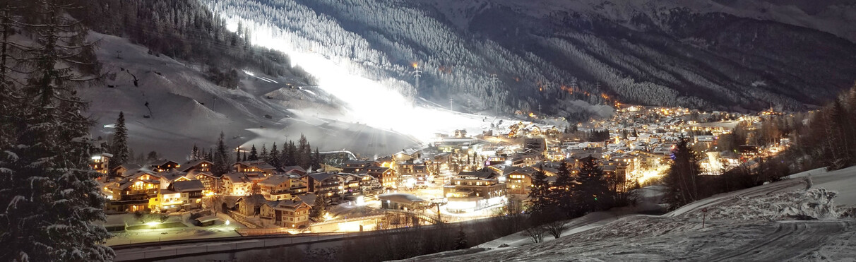 Drohnenaufnahmen von einem verschneiten, winterlichen St. Anton am Arlberg bei Nacht. Umgeben von einer schneebedeckten Bergkette.