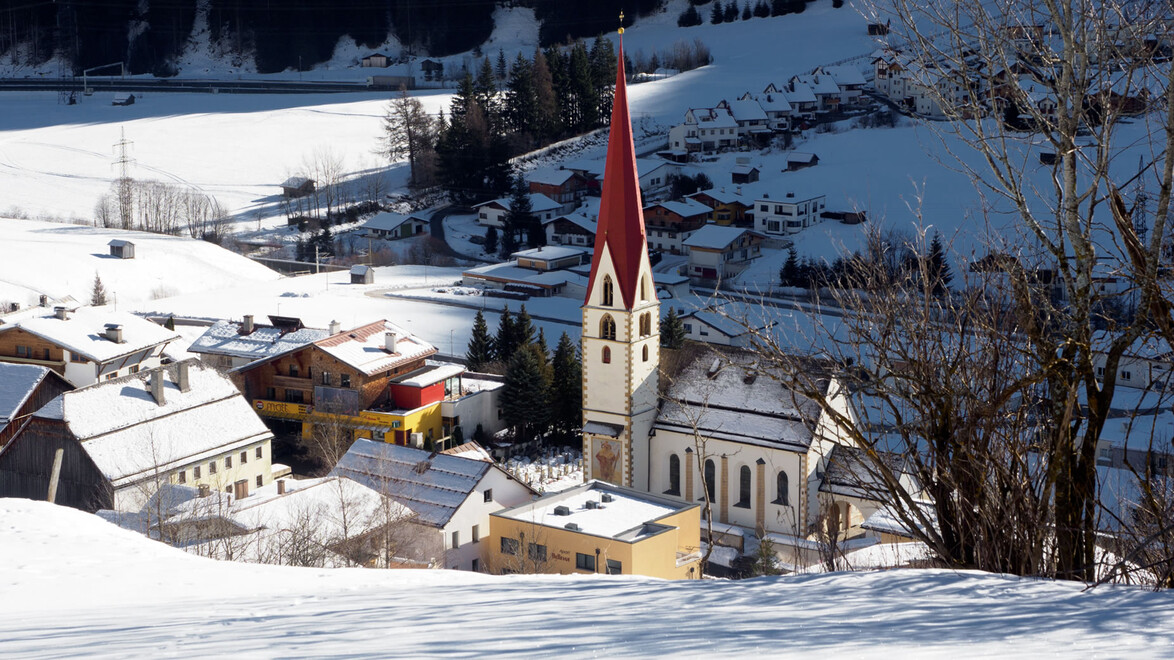 Die verschneite Pettneu Kirche im Fokus einer Aufnahme von etwas höher am Berg /  The snow-covered Pettneu church in the focus of a shot from a little higher up the mountain