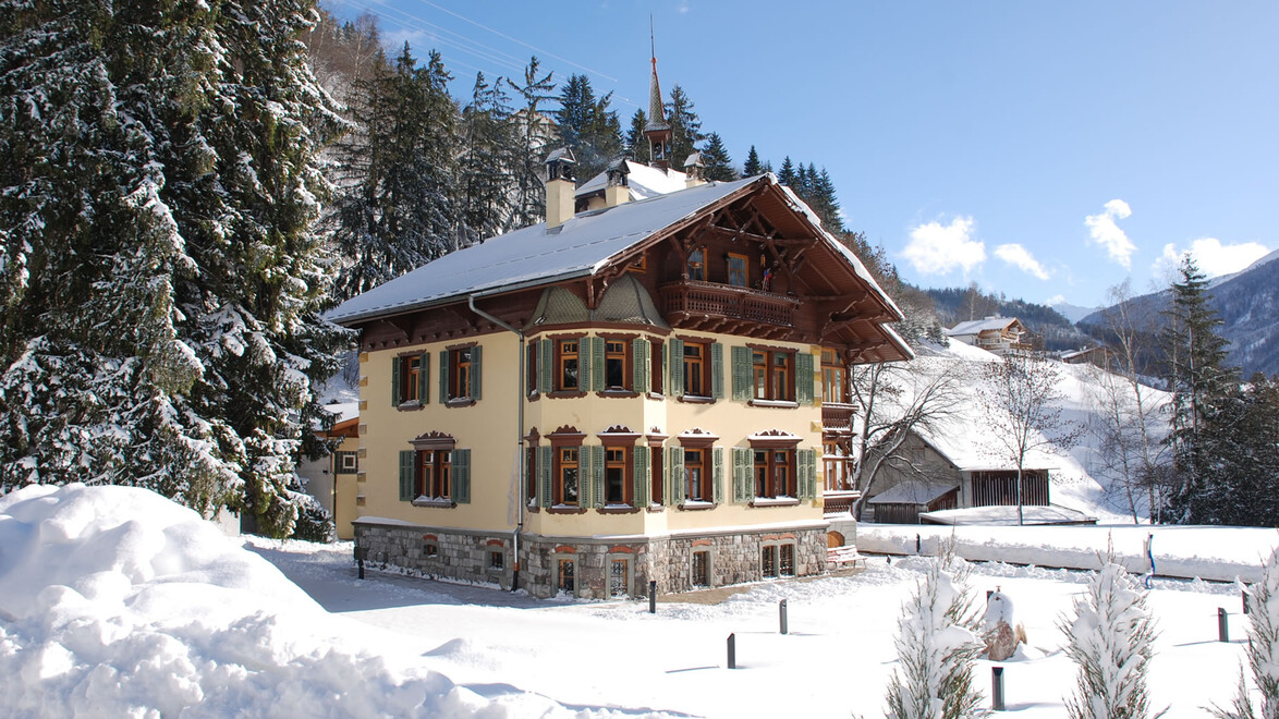 Historisches Gebäude im Schnee mit Bergkulisse im Hintergrund. / Historic house surrounded by snow and forest with mountain backdrop.